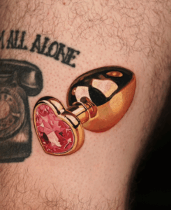 Tattoo of a gold metallic butt plug with a pink heart-shaped gem, showcasing unique body art style.